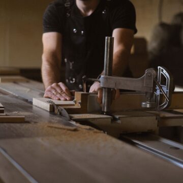 Anonymous male woodworker in apron standing at sawbench while working with wooden details in professional carpentry with stack of wood