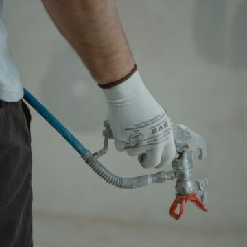 Arm of a worker holding a spray gun wearing gloves in an indoor setting, ready for painting.
